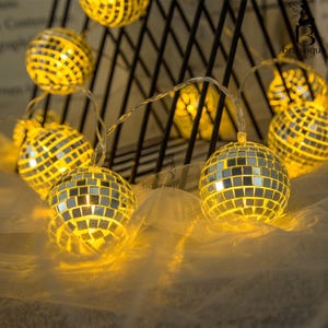 Può includere: Una stringa di luci a sfera da discoteca illuminate. Ogni sfera è ricoperta da piccoli quadrati riflettenti, che emettono un bagliore caldo e giallo. Le luci sono infilate su un filo sottile e argentato, creando un'atmosfera festosa e decorativa.