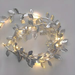 May include: A decorative string of lights featuring metallic silver leaves arranged along a thin wire. The strand is coiled into a circular shape, with small, warm-toned LED bulbs interspersed among the textured, serrated silver foliage.