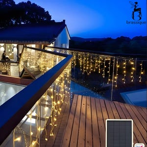 May include: A set of warm white LED icicle string lights draped over a glass balcony railing at night. The lights are powered by a small, rectangular black solar panel shown in the foreground. The logo for brassique is in the top right corner.