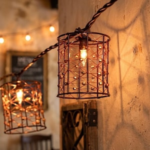 May include: A string of decorative lights featuring cylindrical cages made of thin, dark copper-colored wire. Each cage houses a small, warm-glowing light bulb. The wire structure creates a geometric pattern, casting intricate shadows on the wall.