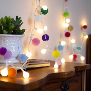 以下が含まれることがあります： 白熱電球と紫、ピンク、青、緑、黄色、オレンジ色のカラフルなポンポンが付いたストリングライト。木製のナイトスタンドの上に、多肉植物が入った白い鉢と開いた本が置かれています。