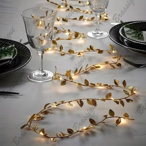 Puede incluir: Guirnalda de hojas doradas con luces LED blancas cálidas. La guirnalda está enhebrada en una mesa blanca con dos copas de vino.