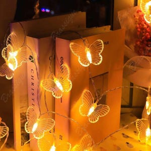 Op de afbeelding: Een lichtsnoer met LED-lampjes voorzien van doorschijnende, vlindervormige kapjes. Elke vlinder wordt van binnenuit verlicht door een warm wit licht, wat een gloeiend effect geeft. De lampjes zijn verbonden door een dunne, flexibele draad.