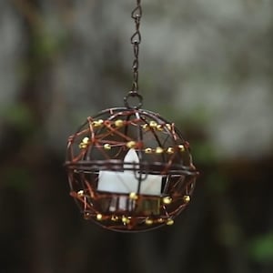 Puede incluir: Un farol esférico colgante construido con alambre marrón oscuro, con un diseño de jaula geométrica acentuado con pequeñas cuentas doradas. Una vela de té blanca se asienta de forma segura dentro del marco de alambre suspendido por una cadena.