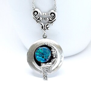 May include: Silver locket necklace with a crescent moon design featuring a woman sitting on the moon. The locket has a blue and green iridescent glass cabochon.