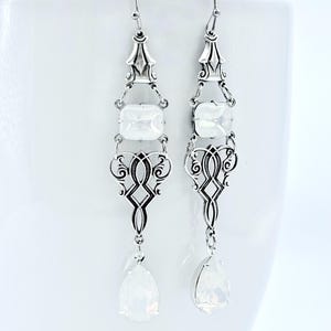 May include: A pair of silver-toned drop earrings with an ornate, filigree design. Each earring features three rectangular white stones and a pear-shaped, opalescent drop. The earrings hang from a simple hook.