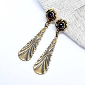 May include: A pair of gold tone dangle earrings with a geometric design and a dark red gemstone in the center of each earring.