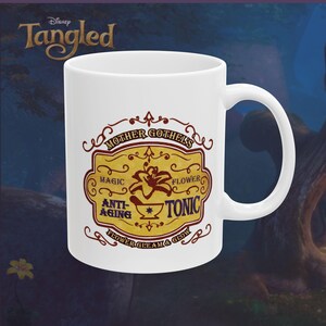 May include: White ceramic mug featuring a design from Disney's Tangled. The mug has a handle and a decorative label with the text "Mother Gothel's Magic Flower Anti-Aging Tonic". The label is in shades of brown and gold.
