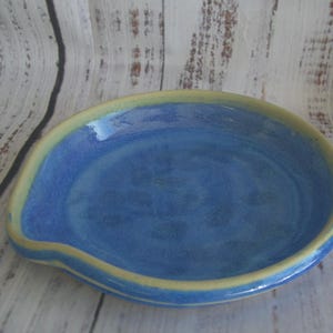 May include: A round ceramic spoon rest with a glossy blue interior and a light yellow rim. The dish features a small indentation on one side, creating a functional shape for holding kitchen utensils while cooking on a stovetop or counter.