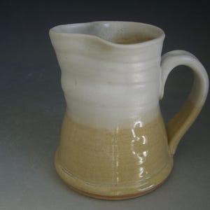 May include: A white and beige ceramic pitcher with a rounded handle. The pitcher has a smooth, glazed finish and a slightly textured base.