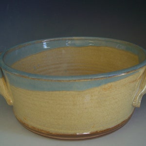 May include: A handmade ceramic bowl with a light yellow body and a blue rim. The bowl has two handles and a brown base. The interior is light yellow. The bowl is approximately 10 inches in diameter.