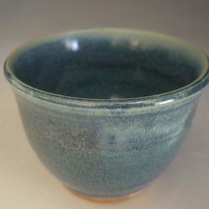 May include: A handmade ceramic bowl with a speckled blue-green glaze. The bowl has a rounded shape with a slightly flared rim and a light brown base. The interior is a darker shade of blue-green.