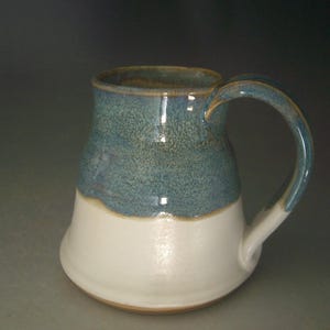May include: A handmade ceramic mug with a two-tone design. The upper portion is a speckled teal, transitioning to a creamy white base. The handle mirrors the teal color. This stoneware mug is ideal for coffee or tea.