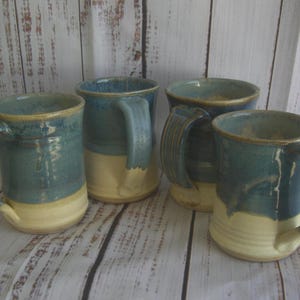 May include: A set of four ceramic mugs featuring a two-tone design with a glossy blue glaze on the upper portion and a matte cream-colored base. Each mug has a sturdy, curved handle and a cylindrical body with subtle horizontal ridges near the bottom.