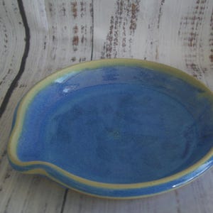 May include: A round ceramic spoon rest featuring a vibrant blue glazed interior and a contrasting pale yellow rim. The piece has a slightly raised edge and a small indentation on one side, showcasing a smooth, glossy finish throughout the surface.