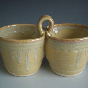 May include: A ceramic serving dish with two bowls and a handle. The bowls are a light yellow color with a brown rim and a glaze that drips down the sides.