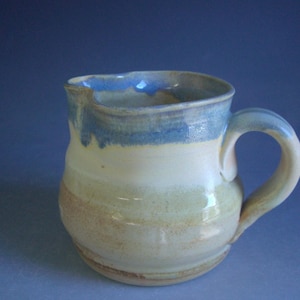 May include: A ceramic pitcher with a light brown, yellow, and green glaze. The pitcher has a blue rim and a handle.