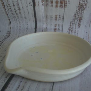 May include: A round, off-white ceramic spoon rest with a shallow basin and a small, integrated spout on one side. The interior surface features subtle, light yellow and blue speckled accents against the smooth, glazed off-white ceramic finish.