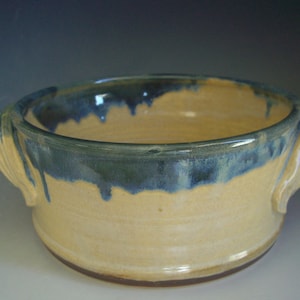 May include: A handmade ceramic casserole dish with two handles. The dish is glazed in a light yellow color with a blue glaze around the rim and handles.