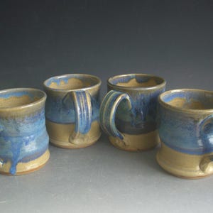 May include: Four handmade ceramic mugs. Each mug has a light tan base, a blue upper section, and a tan rim. The mugs have large, curved handles and a glossy finish. The mugs are arranged in a group.