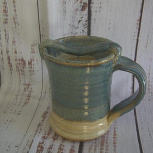 May include: A ceramic mug featuring a two-toned glaze with a dusty blue upper section and a cream-colored base. The mug has a curved handle and a unique horizontal bar extending across the top opening, connecting to the handle and the rim.