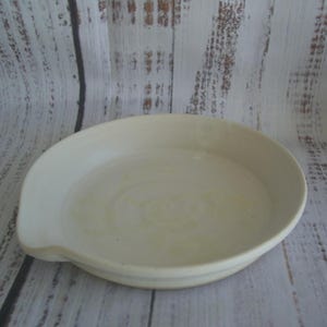May include: A round, off-white ceramic spoon rest with a smooth, glazed finish and a slightly raised edge. The dish features a small, integrated notch on one side, designed to hold the handle of a kitchen utensil securely in place.