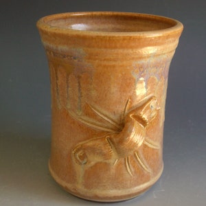 May include: A ceramic mug with a light brown glaze and a raised design of a stylized animal. The mug has a slightly iridescent finish.