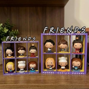 Può includere: Due scaffali viola e gialli con il testo "FRIENDS" in alto, ciascuno con sei statuette di personaggi. Le statuette sono dei personaggi della serie TV "Friends". Gli scaffali sono su una superficie di legno.
