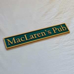 Könnte beinhalten: Ein rechteckiges Schild mit dem Text "MacLaren's Pub" in goldfarbenen Buchstaben. Das Schild hat einen dunkelgrünen Hintergrund und einen goldenen Rand. Das Schild ist wahrscheinlich für einen Pub oder eine Bar.