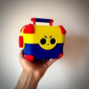 Può includere: Una scatola colorata con manico, con parte superiore gialla, base blu e dettagli rossi. La scatola ha una faccia da cartone animato con occhi neri e un'espressione severa. Il manico è blu.