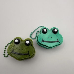 Pode incluir: Dois chaveiros em forma de sapo em tons de verde. Os chaveiros têm olhos pretos e uma boca sorridente. Cada chaveiro tem uma corrente verde correspondente. Os chaveiros são feitos de um material texturizado.