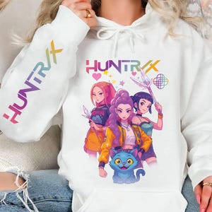 Peut inclure: Un sweat à capuche blanc avec le mot "HUNTRIX" en dégradé arc-en-ciel sur la manche et la poitrine. Le graphisme de la poitrine représente trois femmes de style anime et un chat bleu. Les femmes portent des tenues colorées et tiennent des armes fantastiques.