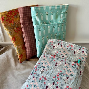 May include: Four fabric book sleeves in various floral and patterned designs. One sleeve is mustard yellow with red flowers, another is brown plaid, a third is teal with white vases and flowers, and the last is light blue with pink flowers.