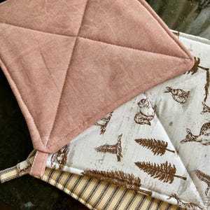 May include: A set of three square quilted pot holders. One is solid dusty rose, one features a white background with brown illustrations of squirrels, deer, and pine trees, and the third has a cream and black vertical striped pattern.