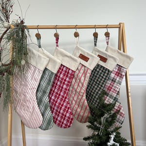May include: Six Christmas stockings hanging on a wooden rack. Each stocking features a cream-colored cuff and a unique fabric pattern, including red and white stripes, green and white plaid, red and white plaid, and green and black plaid.