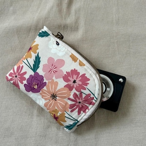May include: A floral print zippered pouch with a black camera partially visible. The pouch features a cream background with pink, purple, and orange flowers. The zipper is a bronze color.