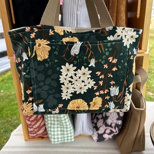 May include: A dark green tote bag with a floral pattern featuring white and yellow flowers. The bag has a front pocket and tan handles. Other bags with different patterns are visible in the background.
