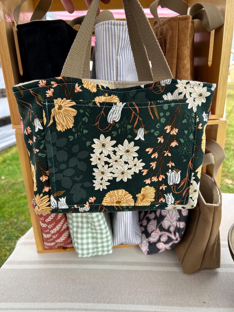 May include: A dark green tote bag with a floral pattern featuring white and yellow flowers. The bag has a front pocket and tan handles. Other bags with different patterns are visible in the background.