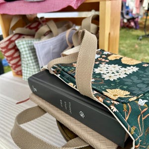 May include: A green floral print Bible cover with a tan handle rests on a table, with a Bible inside. The cover has a white interior and a tan strap. Other tote bags are visible in the background.
