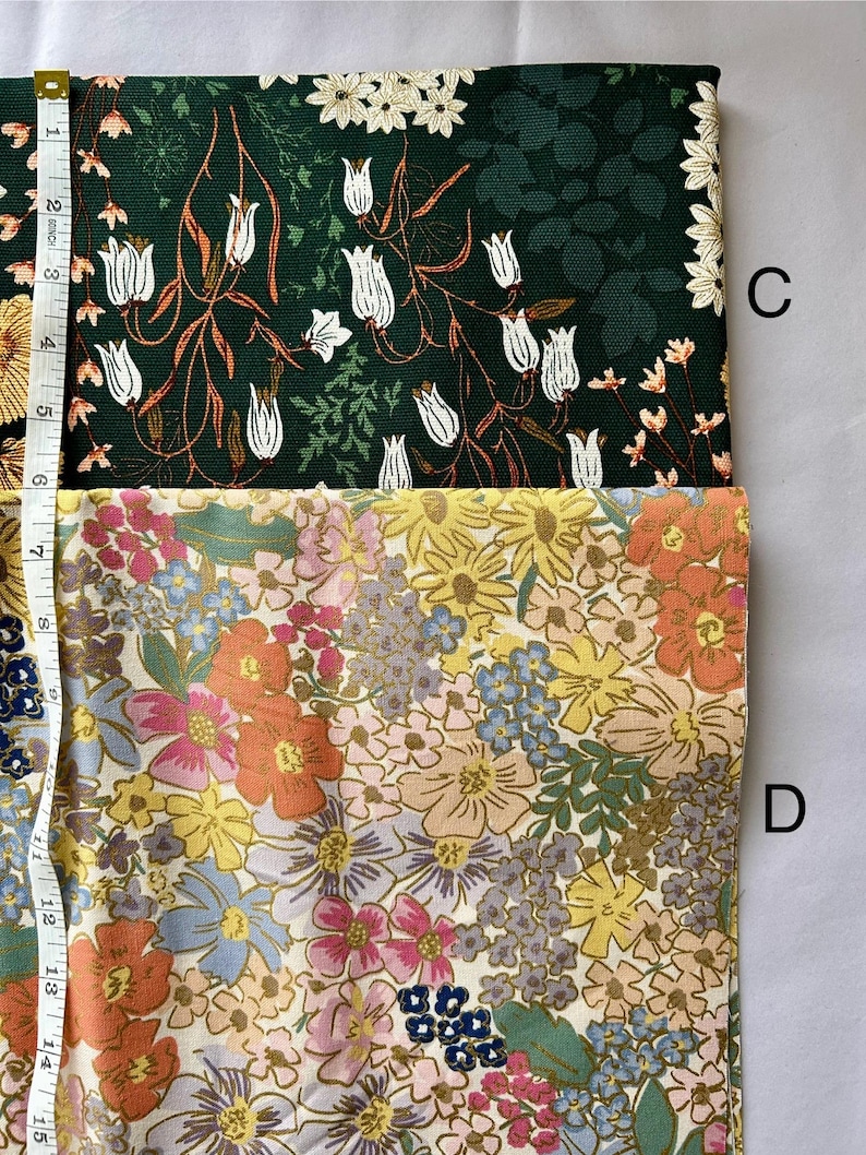 May include: Two fabric swatches with floral patterns. One is dark green with white tulip-like flowers and orange stems. The other is a mix of colorful flowers in shades of yellow, orange, pink, and blue. A measuring tape is visible.