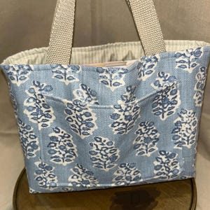 May include: A small tote bag with a light blue floral pattern and a front pocket. The bag has beige handles and a beige interior. The floral design is white and blue. The bag is sitting on a wooden surface.