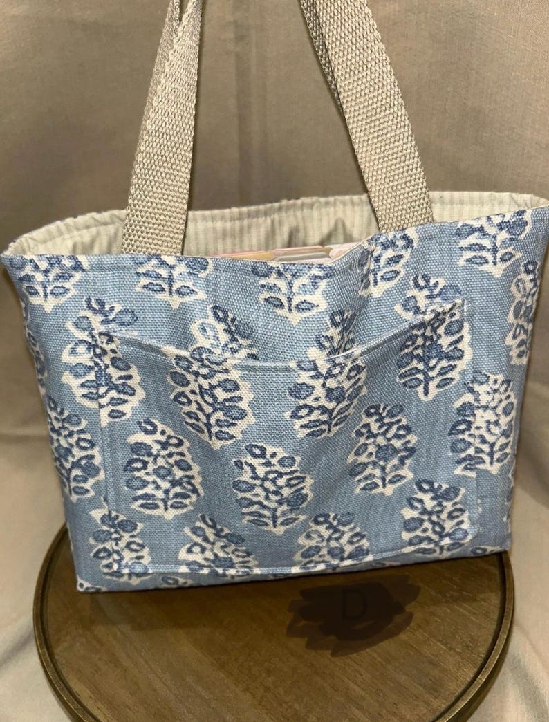May include: A small tote bag with a light blue floral pattern and a front pocket. The bag has beige handles and a beige interior. The floral design is white and blue. The bag is sitting on a wooden surface.