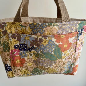 May include: A floral print tote bag with a beige interior and brown handles. The bag features a colorful pattern of flowers in shades of pink, yellow, blue, and orange. The tote has a small pocket on the front.