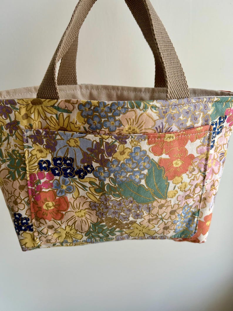 May include: A floral print tote bag with a beige interior and brown handles. The bag features a colorful pattern of flowers in shades of pink, yellow, blue, and orange. The tote has a small pocket on the front.