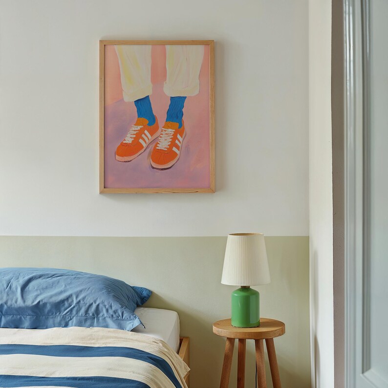 May include: A framed painting of a pair of orange sneakers with blue socks and white pants on a pink background. The painting is hanging on a white wall above a bed with blue and white striped bedding.