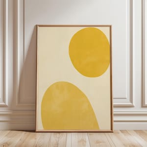 Puede incluir: Una impresión artística abstracta enmarcada con dos formas amarillas sobre un fondo crema. La forma superior es un círculo, y la forma inferior es redondeada. La obra de arte se exhibe en una habitación con suelo de madera y paredes blancas.