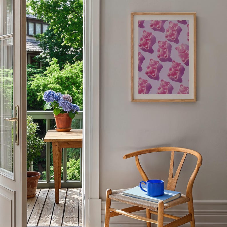 Pode incluir: Uma impress&atilde;o emoldurada com um padr&atilde;o repetido de ursinhos de goma rosa sobre um fundo lil&aacute;s. A impress&atilde;o est&aacute; pendurada em uma parede cinza, ao lado de uma cadeira de madeira com uma caneca azul e um livro. O quarto tem vista para uma varanda com plantas.