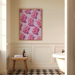 Pode incluir: Uma impress&atilde;o emoldurada com um padr&atilde;o repetitivo de ursinhos de goma rosa sobre um fundo lil&aacute;s claro. A obra de arte &eacute; exibida em um banheiro, adicionando um toque de cor &agrave; decora&ccedil;&atilde;o neutra.