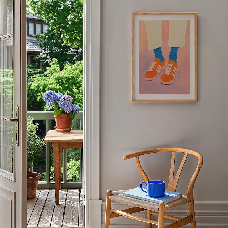 May include: A wooden chair with a woven seat and a blue ceramic mug on top of a book. A framed illustration of a person wearing orange sneakers and blue socks hangs on the wall behind the chair.