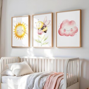 May include: Three framed watercolor art prints featuring a smiling sun, a bee on a flower, and a smiling pink cloud. The prints have a light wood frame and are displayed above a white crib with bedding.